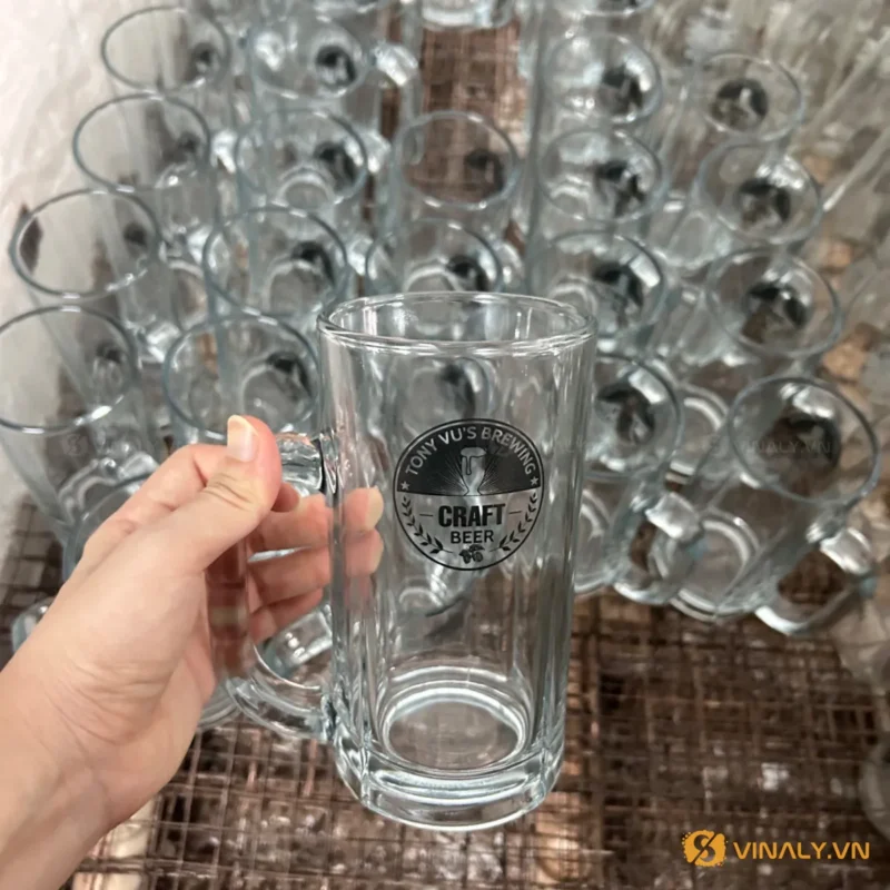 Một bàn tay đang cầm cốc bia thủy tinh in logo đen rõ nét, xung quanh là hàng loạt cốc thủy tinh quai cầm khác tại xưởng