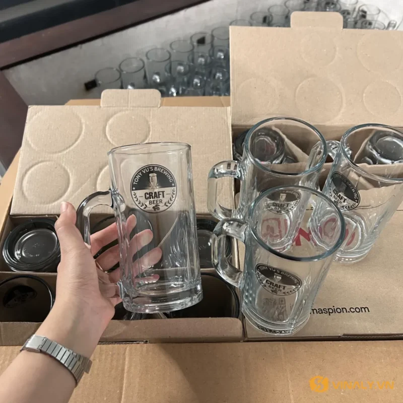 Bàn tay cầm cốc bia thủy tinh in logo, nhiều cốc có quai khác trong thùng carton nâu, sẵn sàng cho đơn hàng số lượng lớn