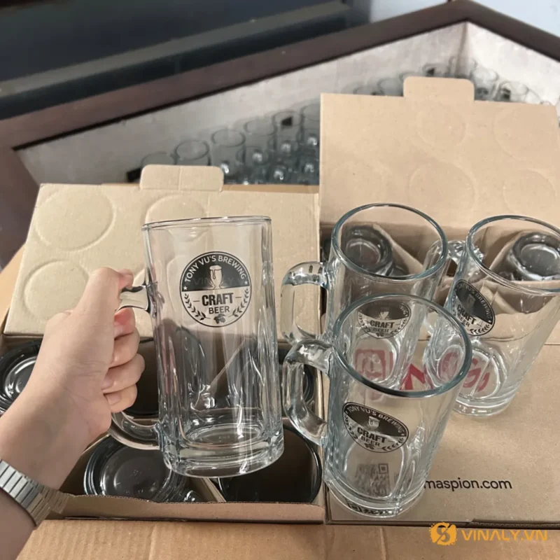 Hình ảnh ly bia thủy tinh trong suốt có quai cầm, được in logo thương hiệu Tony Vu's Brewing, xếp trong hộp carton. Sản