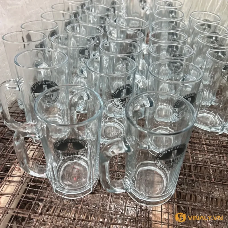 Hàng loạt ly bia thủy tinh trong suốt có quai cầm chắc chắn, một số ly đã được in logo thương hiệu Craft Beer màu đen, đ