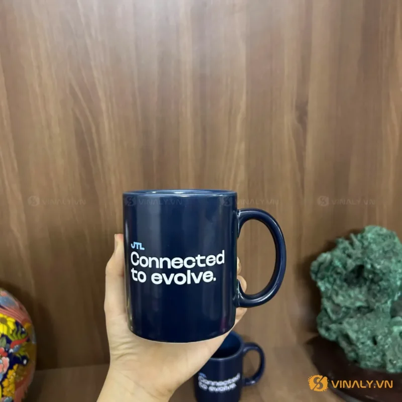 Hình ảnh ly sứ trụ xanh cửu long in logo JTL màu trắng, được một bàn tay cầm trên nền gỗ, phía sau có một ly nhỏ hơn và