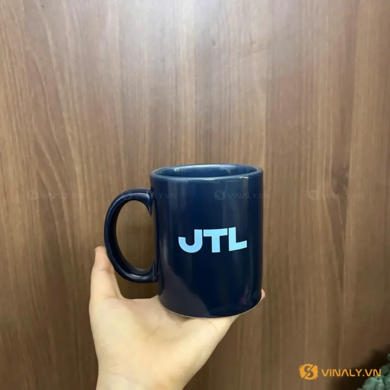Một bàn tay đang cầm ly sứ màu xanh navy in logo JTL màu xanh nhạt, đặt trên nền gỗ. Ly có quai tròn, bề mặt trơn bóng.