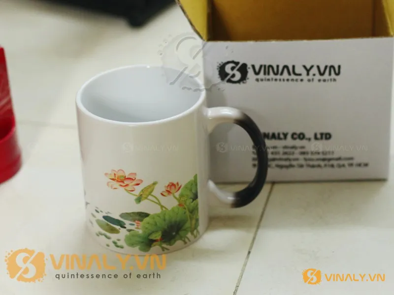 Ly sứ trắng đổi màu in hoa sen tinh tế, quai đen trắng, đặt cạnh hộp quà Vinaly.vn, sẵn sàng in logo doanh nghiệp.