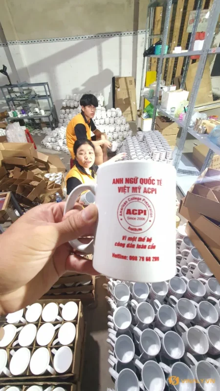 Một người đang cầm ly sứ trắng quai tròn in logo ACPI màu đỏ, phía sau là xưởng sản xuất với hàng trăm ly sứ trắng in logo