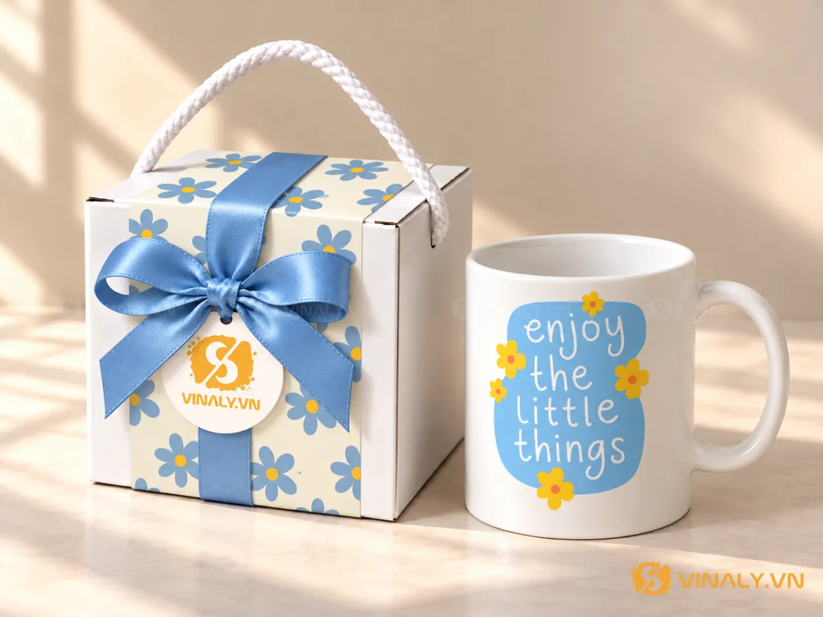 Set Quà Tặng Ly Sứ Cao Cấp Phụ Nữ 20/10 8/3 12 Ly sứ trắng quai tròn in logo với họa tiết chữ và hoa vàng xanh, đặt cạnh hộp quà hoa văn cùng nơ xanh dương đẹp mắt.
