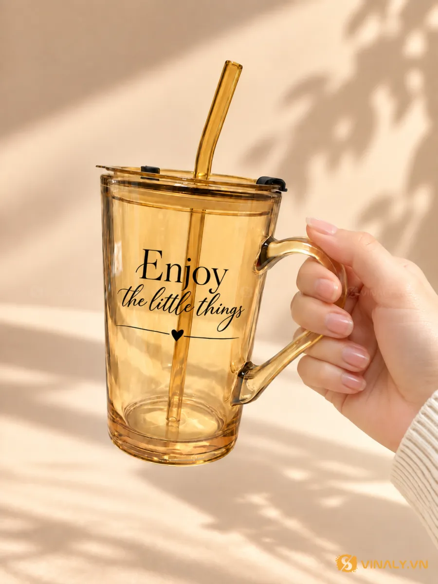 Một bàn tay đang cầm ly thủy tinh màu hổ phách có quai, kèm nắp và ống hút, in chữ 'Enjoy the little things'. Ly thủy tinh