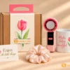 Ảnh chụp set quà tặng ly sứ quạt mini in logo màu hồng và trắng, kèm hộp giấy, dây buộc tóc, thiệp trên nền be nhạt.