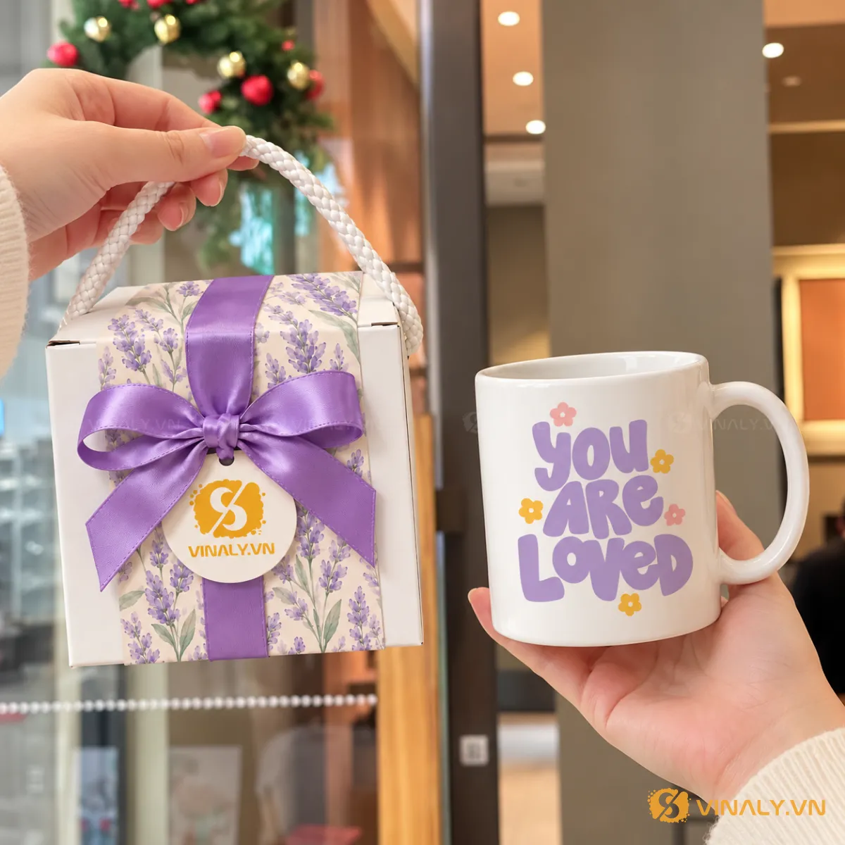 Set Quà Tặng Ly Sứ Cao Cấp Phụ Nữ 20/10 8/3 5 Hình ảnh cận cảnh ly sứ trắng quai tròn với dòng chữ 'You Are Loved' màu tím và hoa vàng, được cầm trên tay, bên cạnh hộp